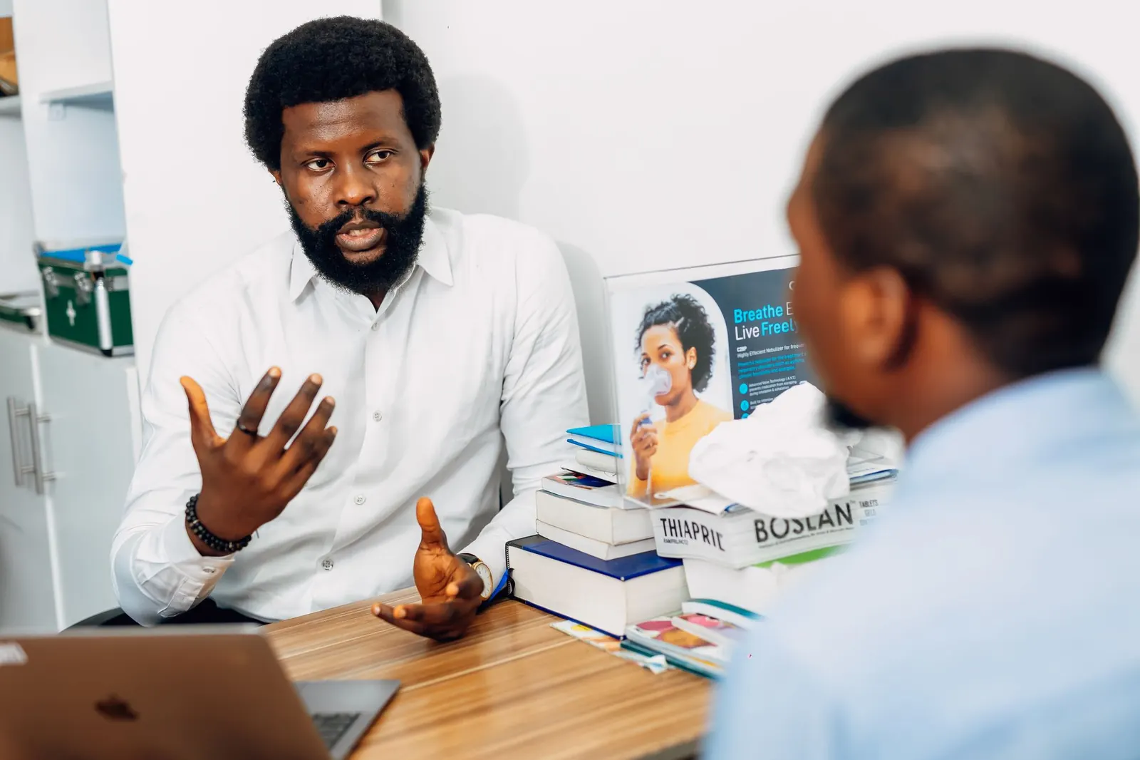 Nigerian doctor in a white shirt explaining a diagnosis to a patient across his desk in a bright consulting room